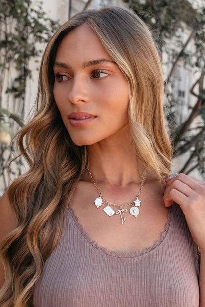 Woman with long wavy hair wears a mauve tank and the Gold Christie Multi Charm Chain Link Necklace outdoors with greenery behind.