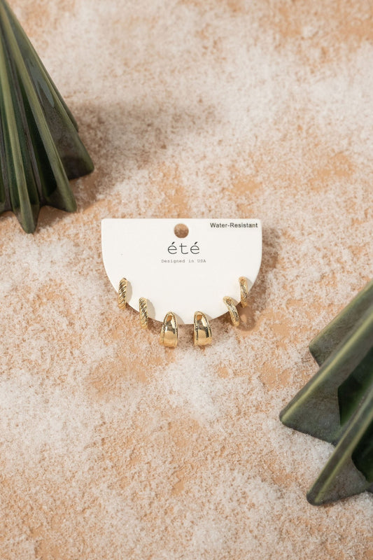 The Gold Chunky Earrings Set on a branded card with green accents rests on sand—stylish, water-resistant, and versatile.
