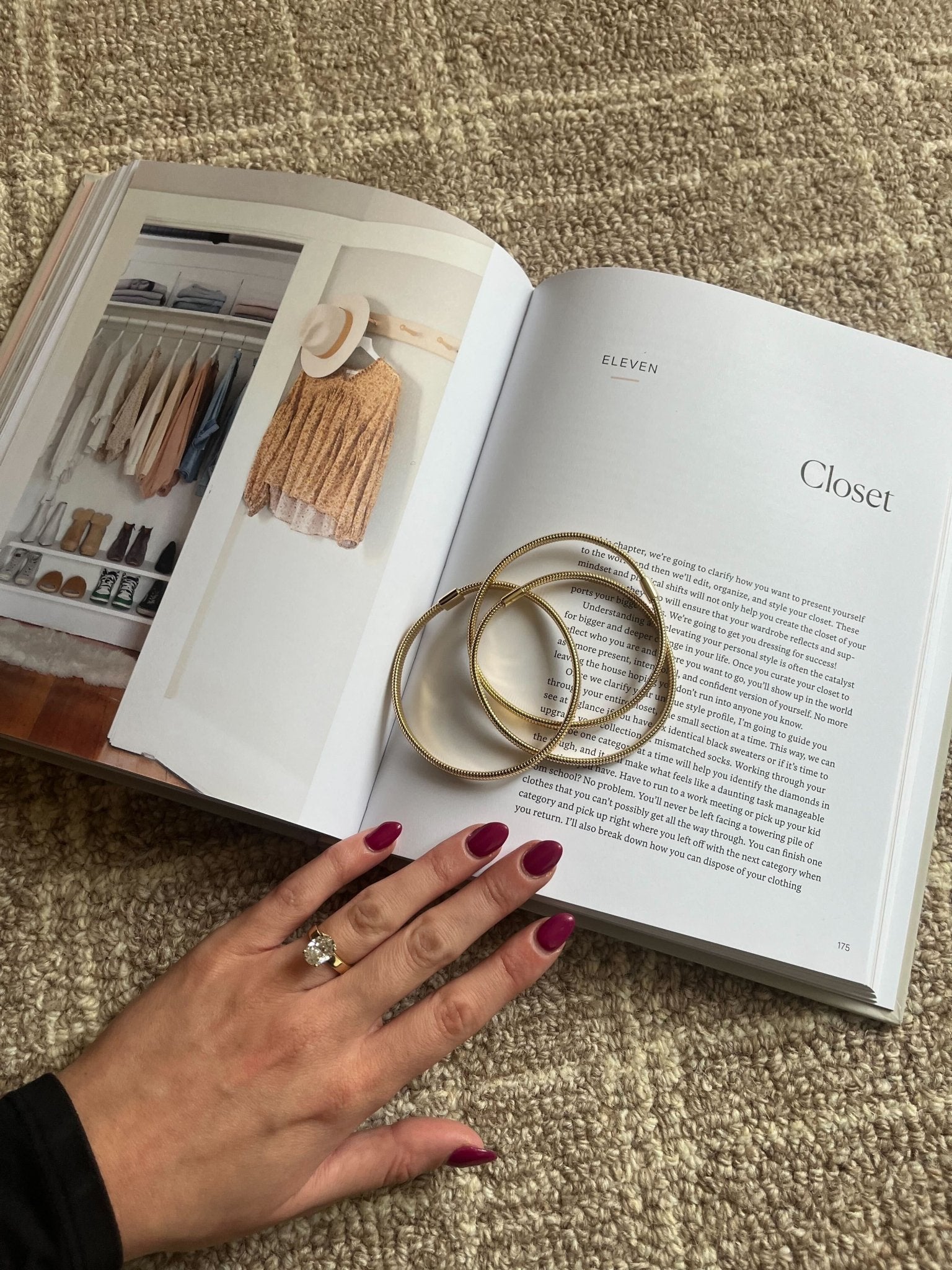 A hand with red nails and a ring wearing the Gold Coiled Metallic Bracelet Set rests on an open book atop a beige carpet.