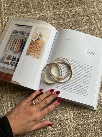 A hand with red nails and a ring wearing the Gold Coiled Metallic Bracelet Set rests on an open book atop a beige carpet.