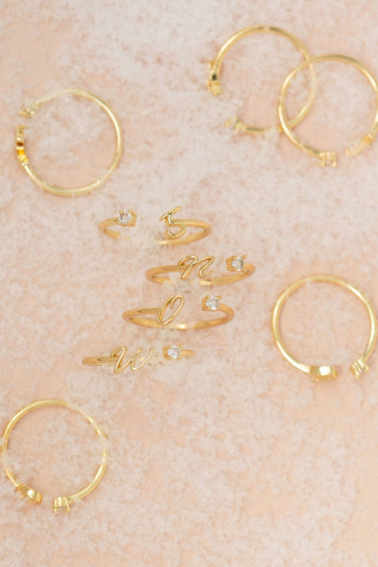 Gold Custom Initial Letter Band Ring with small diamonds, ideal personalized jewelry, shown on a peach-textured background.