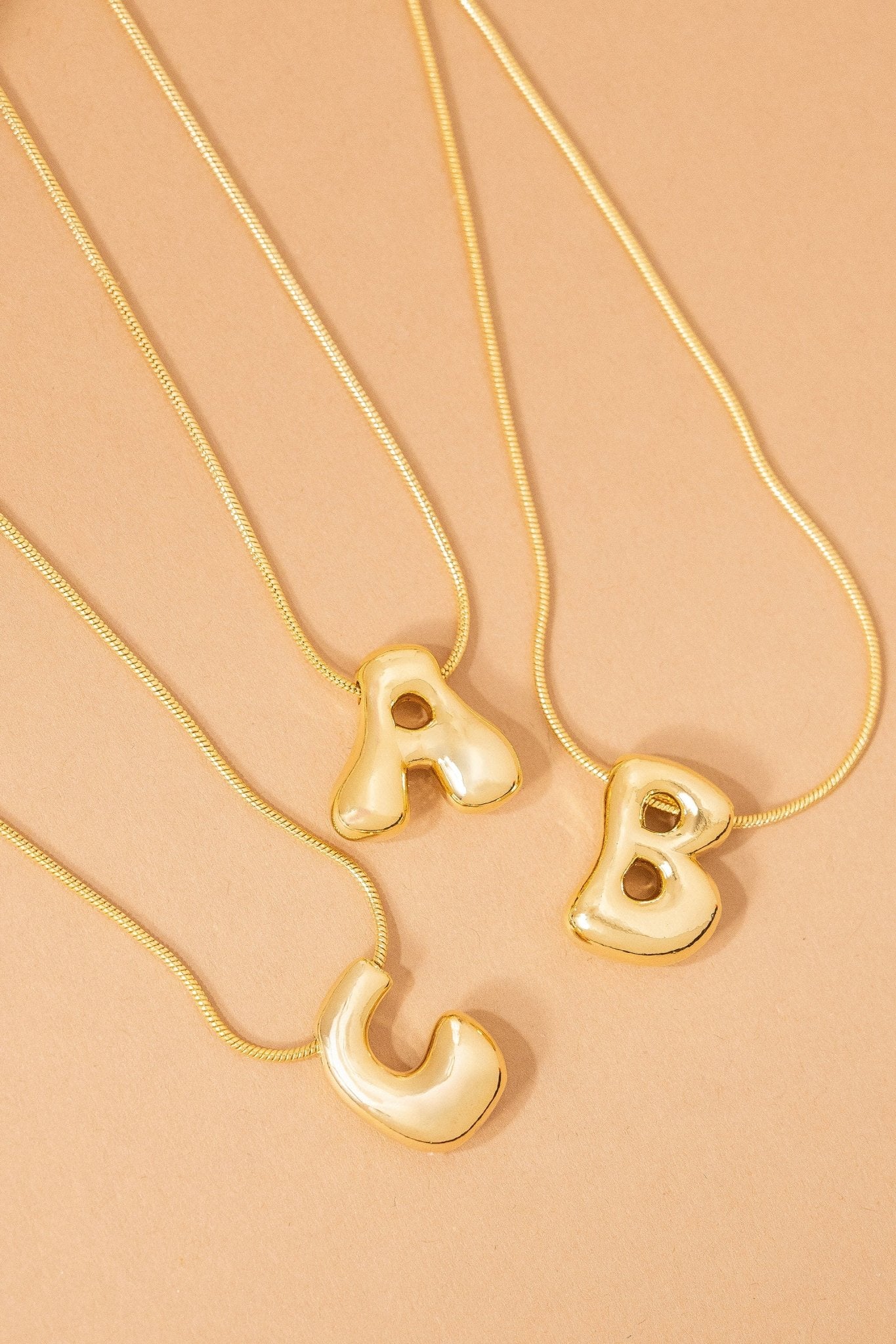 Three Gold Dipped Bubble Letter Pendant Necklaces—A, B, and C—are arranged on a beige surface.