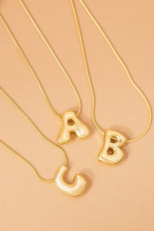 Three Gold Dipped Bubble Letter Pendant Necklaces—A, B, and C—are arranged on a beige surface.