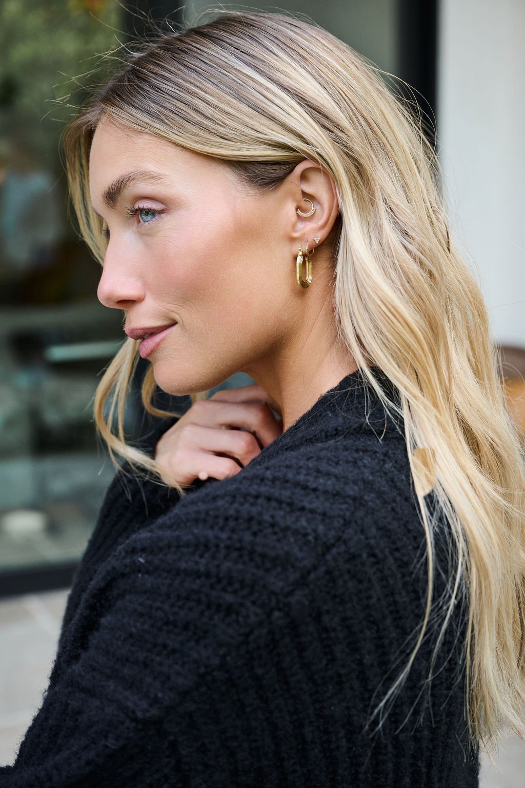 Woman with long blonde hair in a black sweater wears Gold Dipped Flat Grooved Hoop Earrings outdoors, looking to the side.