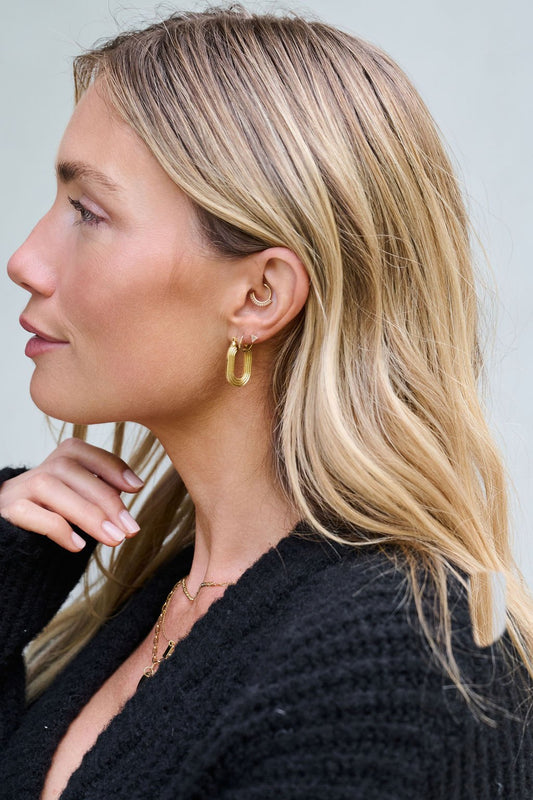 A woman with wavy blonde hair models Gold Dipped Flat Grooved Hoop Earrings, wearing a black textured top in profile.