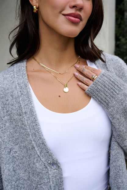 A woman wears the Gold Dipped Pendant Layered Necklace, gold rings, a white top, and a gray cardigan. Her dark hair is shoulder-length.