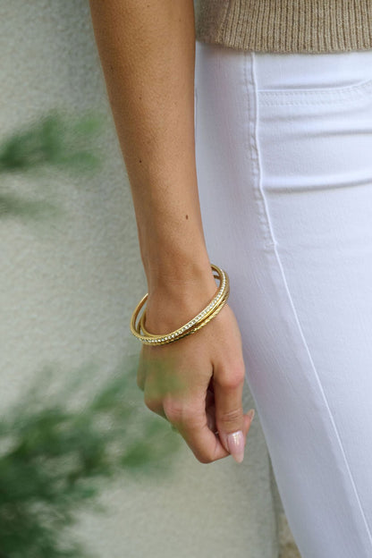 A woman’s arm wearing the Gold Double Layered Bracelet Set, in a tan top and white pants, with a blurred green plant in front.