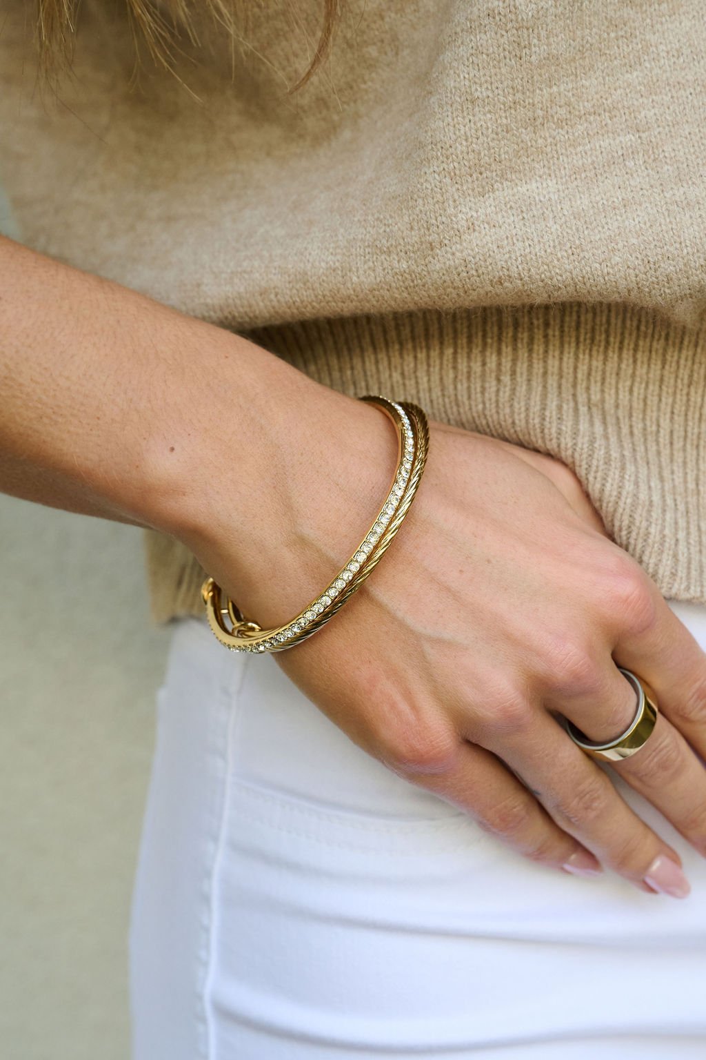Wearing the Gold Double Layered Bracelet Set, a person in a beige sweater and white pants rests their hand on their hip.