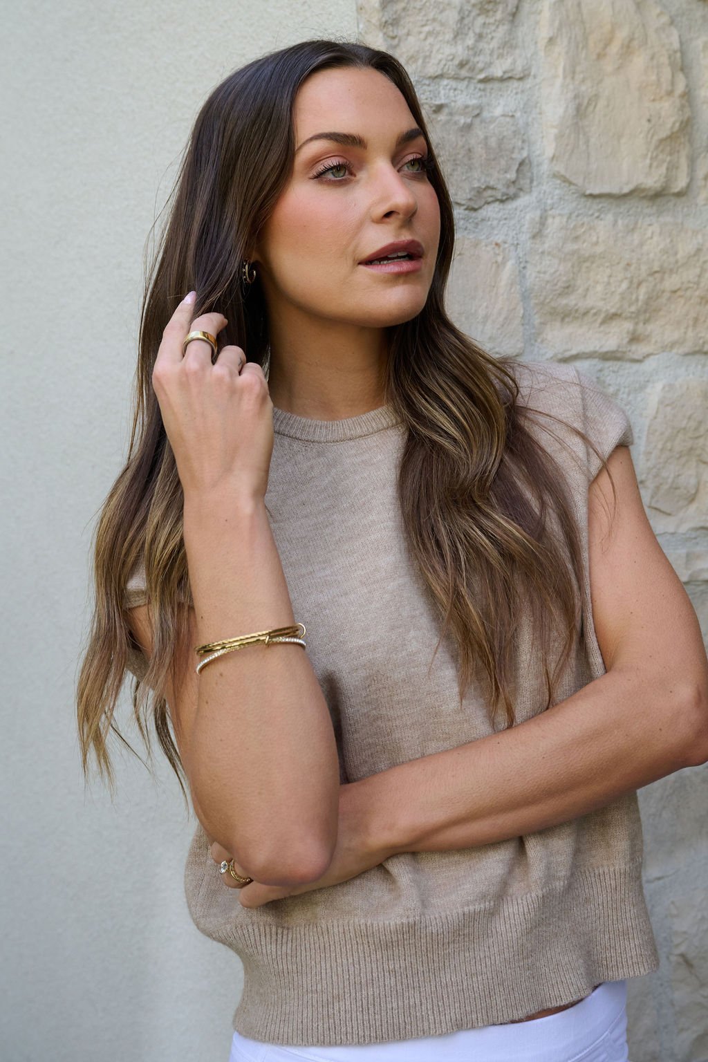 A woman with long brown hair, in a beige sleeveless top and Gold Double Layered Bracelet Set, stands by a light stone wall.