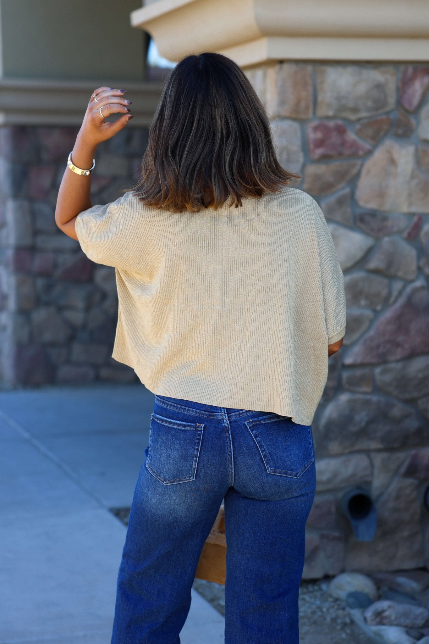 Wearing a Gold Glittery Short Sleeve Top and blue jeans, a person with shoulder-length hair stands outside facing a stone wall, raising their left arm.