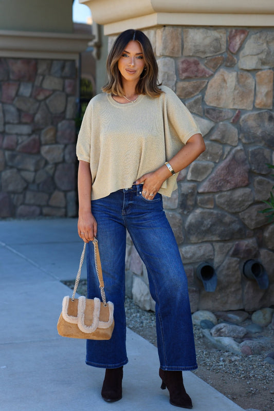 Woman smiles outdoors wearing a Gold Glittery Short Sleeve Top, blue jeans, brown boots, and holding a tan handbag.