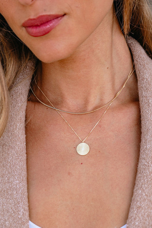 A woman wears the Gold Layered Disc Pendant Necklace over a cream top with a beige textured jacket.