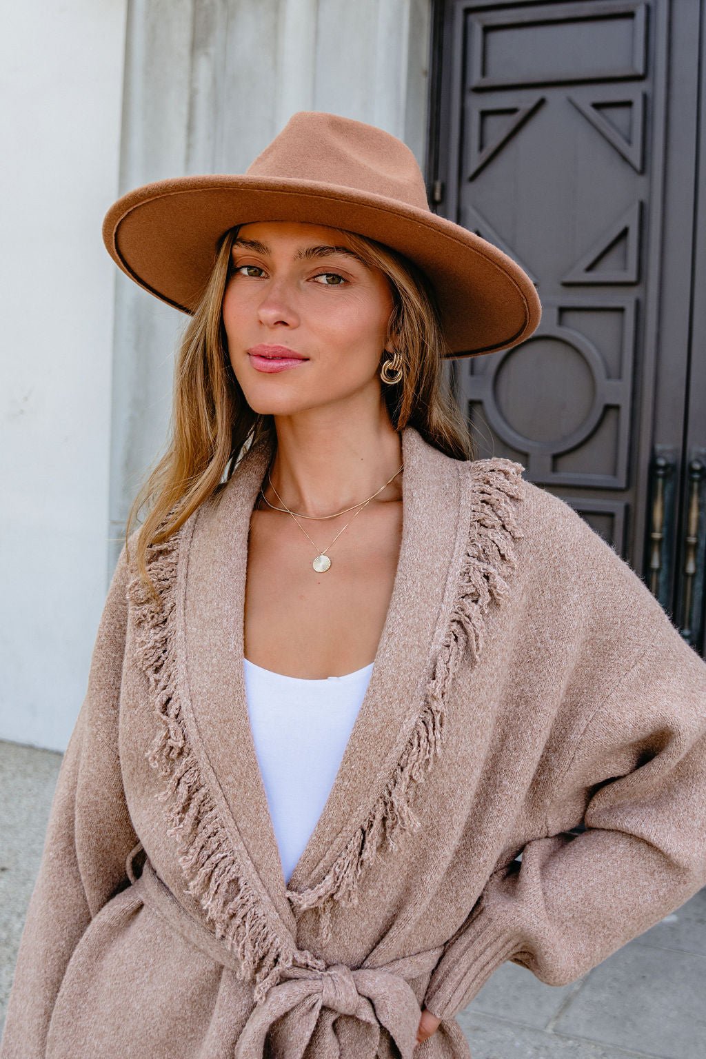 Woman outside in a tan fringed cardigan and hat wears the Gold Layered Disc Pendant Necklace, standing before a decorative door.