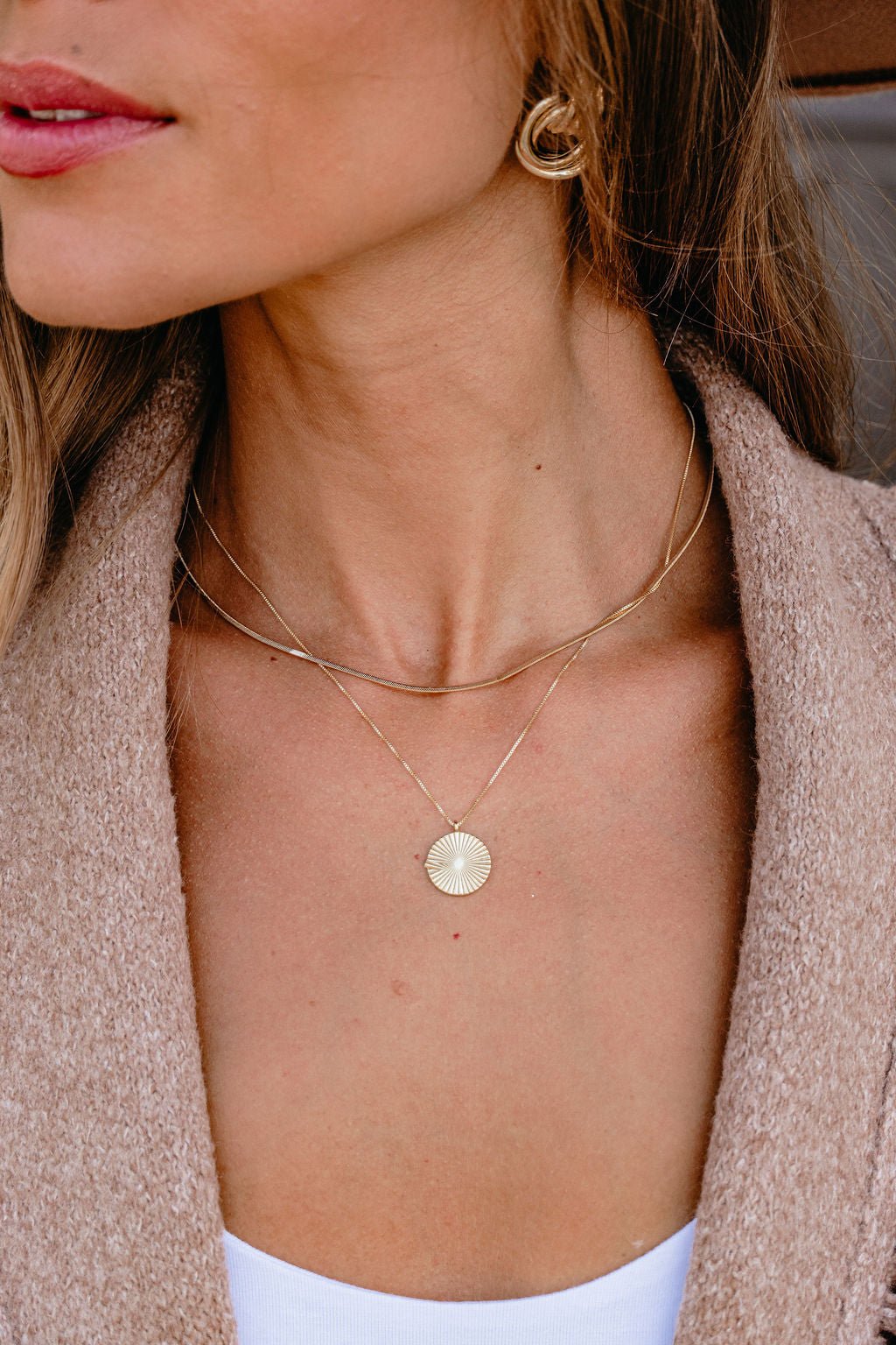 Woman wearing a Gold Layered Disc Pendant Necklace, gold earrings, beige jacket, and white top.