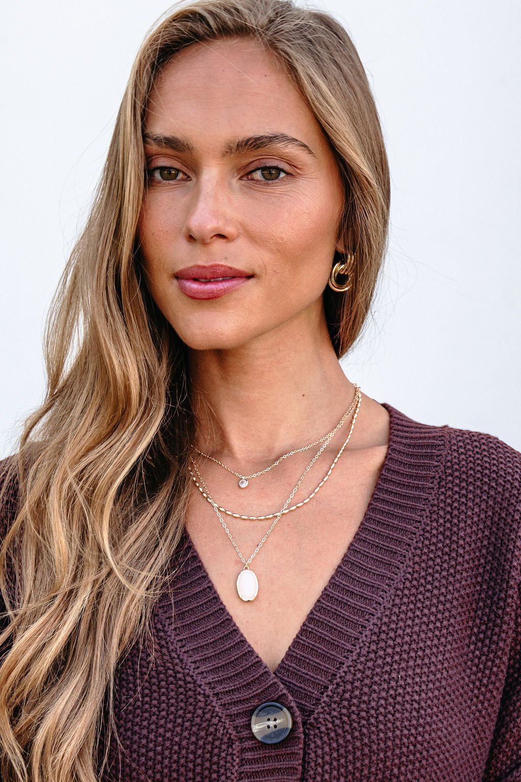 Woman with long blonde hair wears the Gold Layered Natural Stone Necklace, hoop earrings, and a purple knit sweater against a white background.