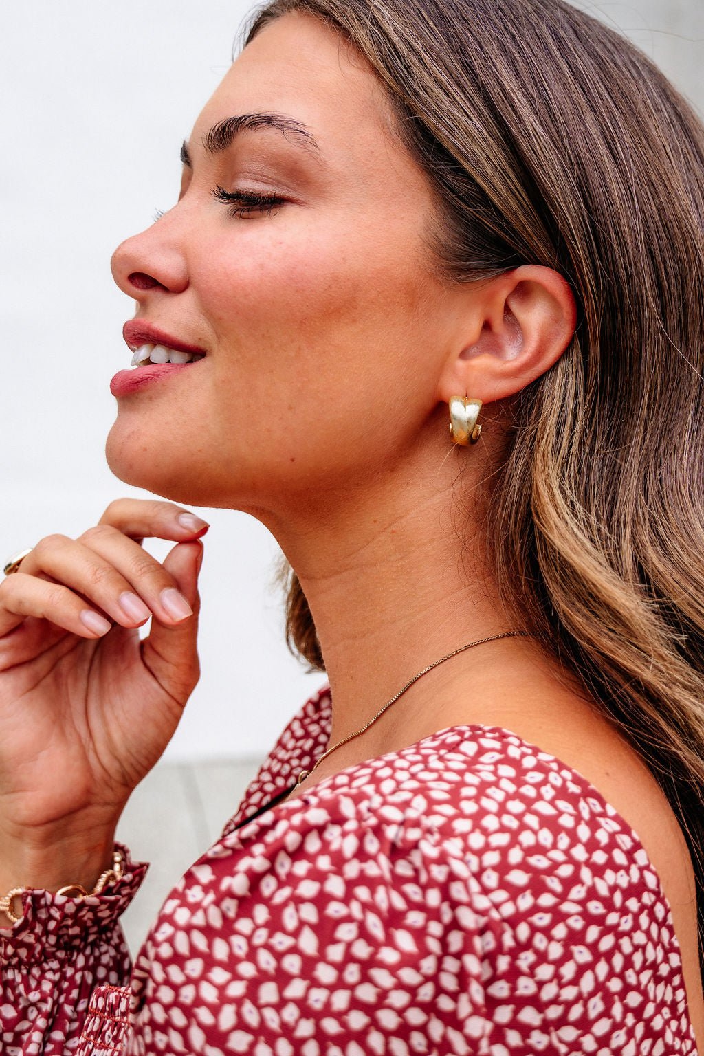 Woman in profile with long brown hair, smiling, wearing Gold Matte Small Hoop Earrings and a red patterned top, touching her chin.
