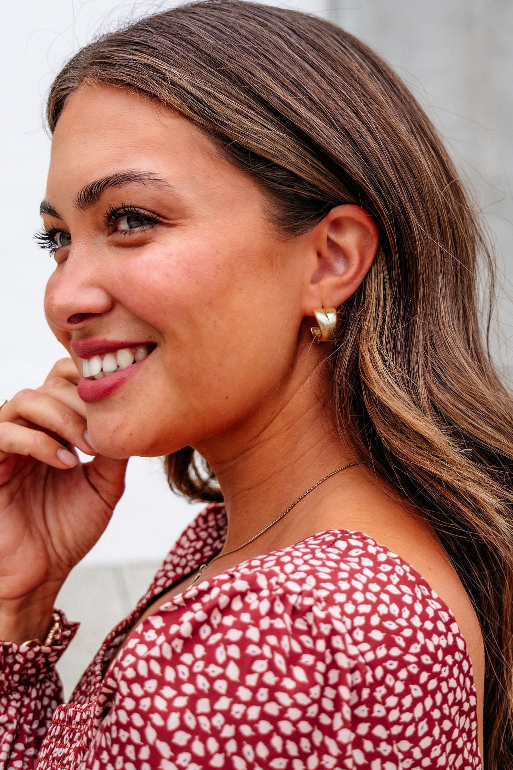 Woman with long brown hair smiles, wearing Gold Matte Small Hoop Earrings and a red patterned top, hand near her face.