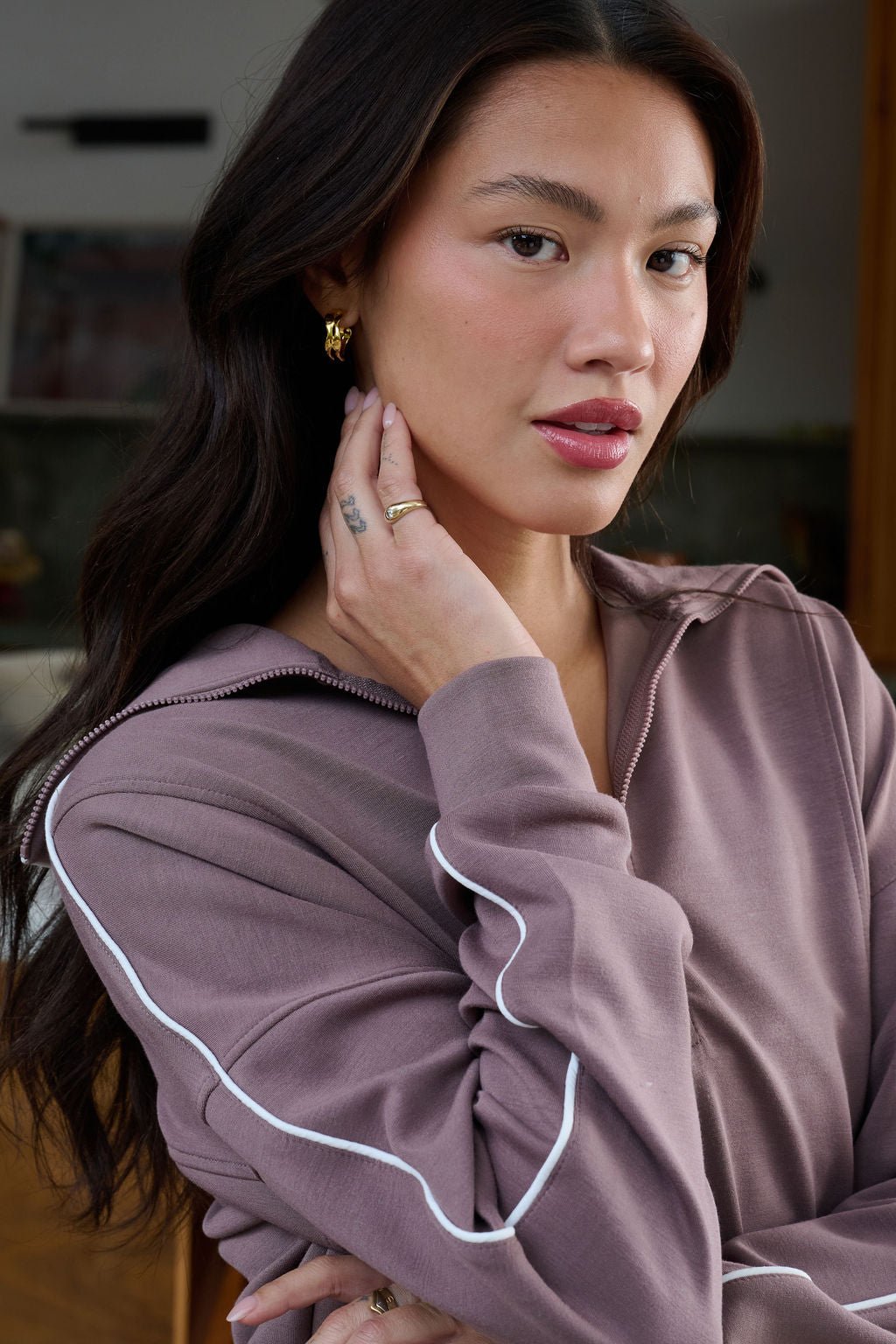 A woman with long dark hair poses indoors, wearing Gold Metallic Open Hoop Earrings and a mauve zip-up top with white sleeve piping.