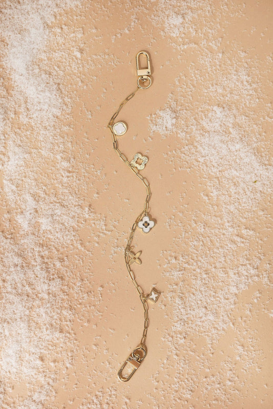 The Gold Multi Clover Charm Purse Chain with floral details rests on a peach surface, perfect as a handbag accessory.