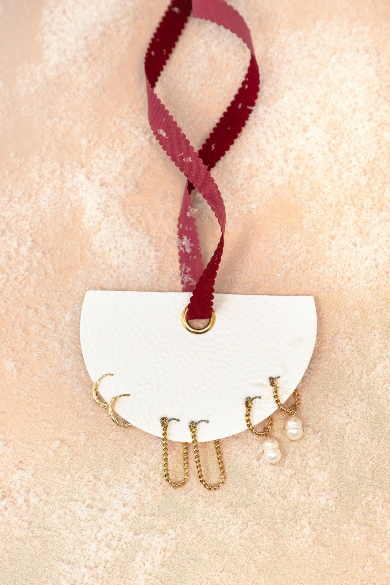 Gold Pearl Charm Twisted Hoop Earrings Set displayed on a white semi-circle holder with a red ribbon on beige textured surface.