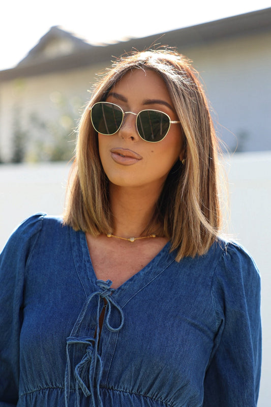 A woman outdoors wears Gold Smoke Lens Narrow Sunglasses, sunlight reflecting off her denim top and straight shoulder-length hair.