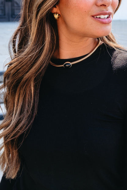 Woman with long wavy hair and black top wears Gold Snake Chain Black Oval Pendant Necklace and gold earrings, smiling slightly.