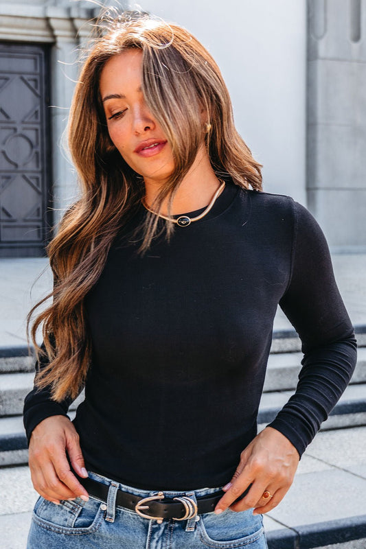 A woman in a black top and jeans smiles on stone steps, wearing the Gold Snake Chain Black Oval Pendant Necklace for a modern touch.