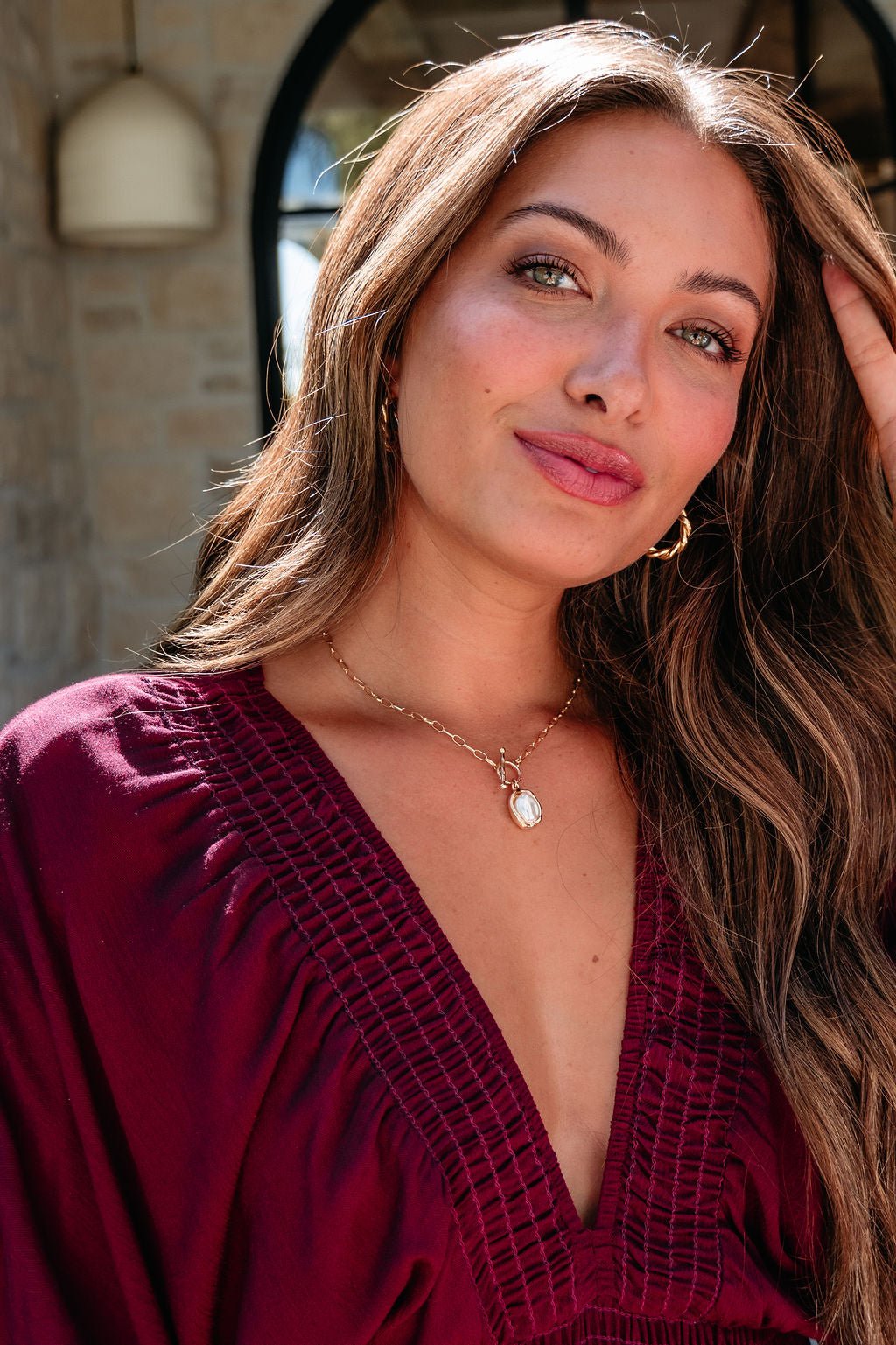 Woman with long brown hair wears a burgundy top and Gold Toggle Chain Pearl Pendant Necklace, smiling softly outdoors.