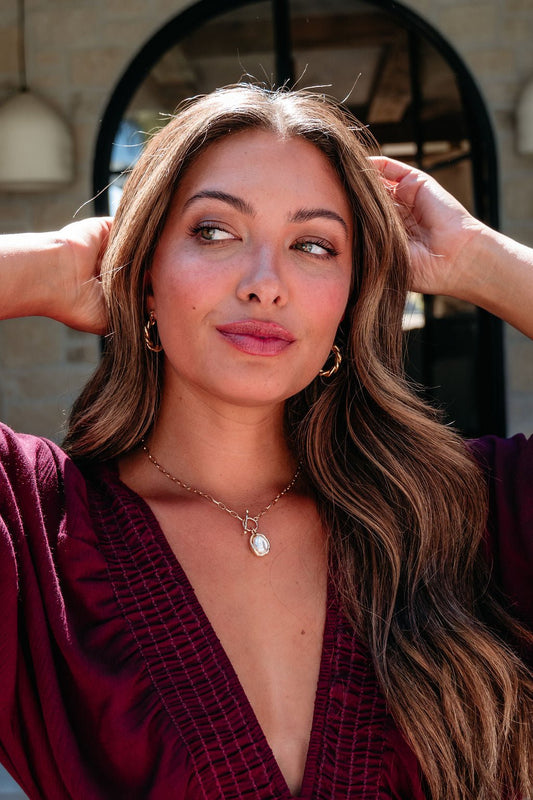 Woman with long wavy hair wears a maroon top and the Gold Toggle Chain Pearl Pendant Necklace, posing outdoors.