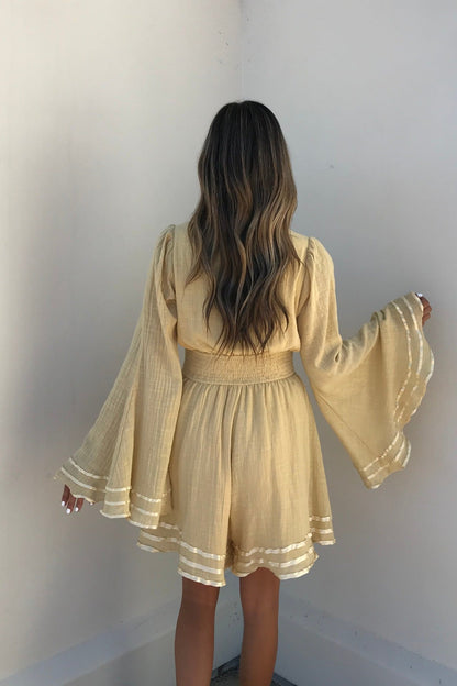 A woman with long wavy hair stands in a corner, wearing the Golden Hour Bell Sleeve Romper featuring wide, flared sleeves.
