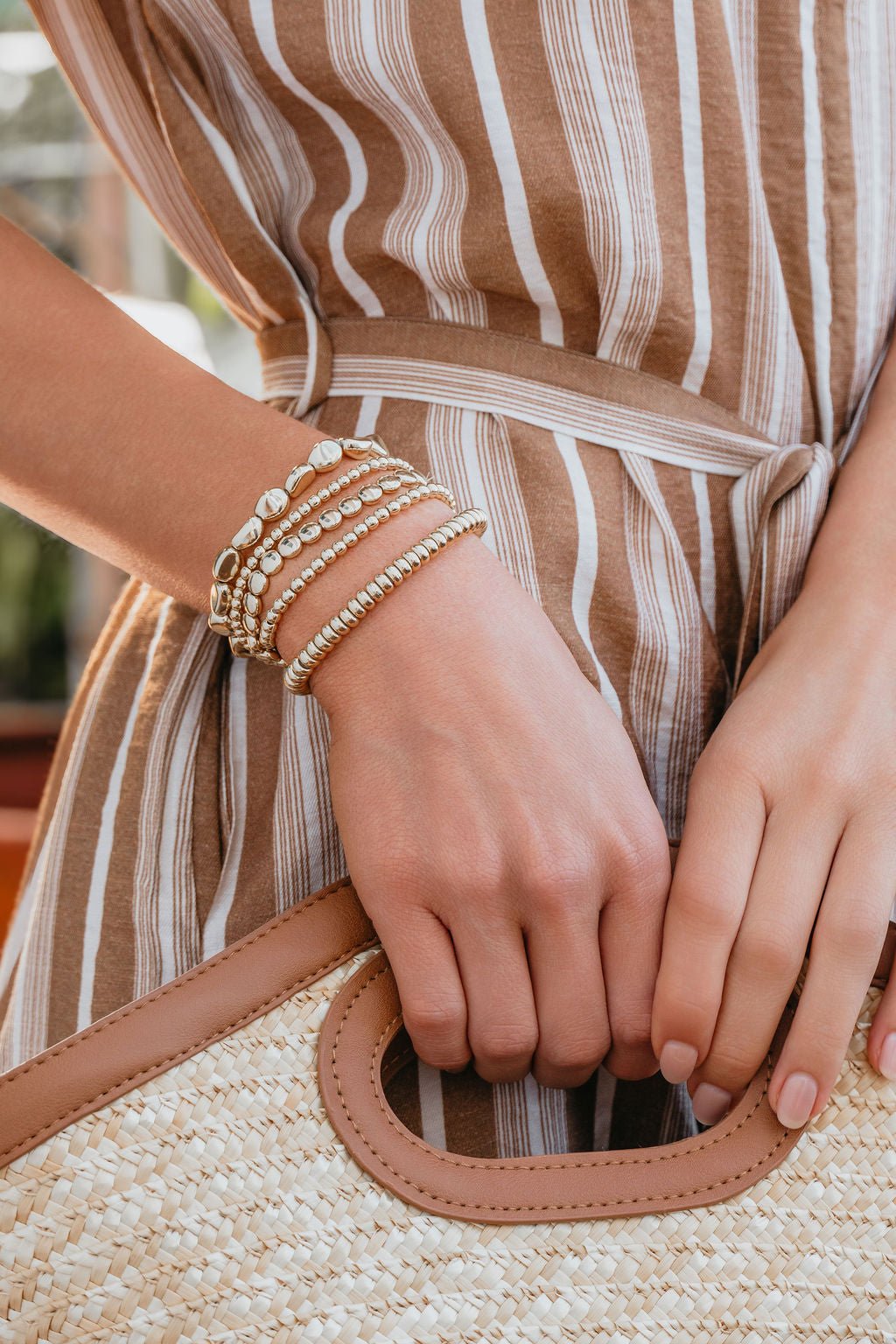 A woman in a brown striped dress holds a straw bag, wearing the Golden Hour Bracelet Set on her wrist.