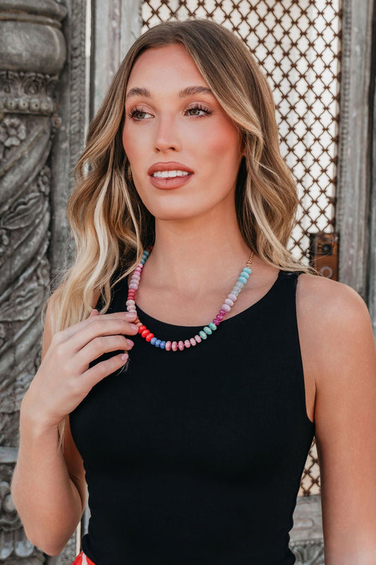 Woman with wavy blonde hair wears a black sleeveless top and the Golden Sol Beaded Necklace, standing by ornate metalwork.