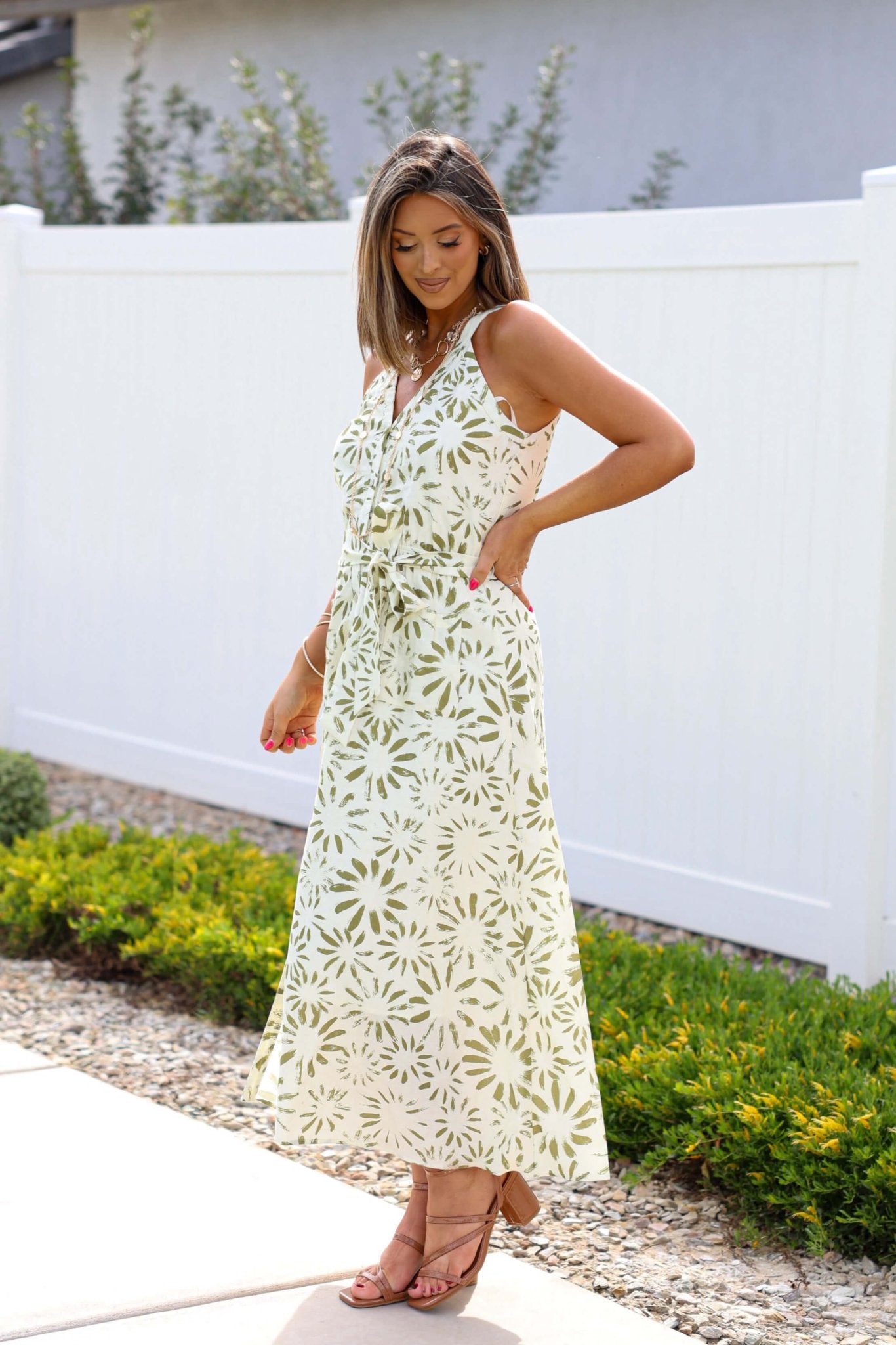 Woman in the Graceful Olive Floral Print Belted Midi Dress stands by a white fence and garden, looking down with a smile.