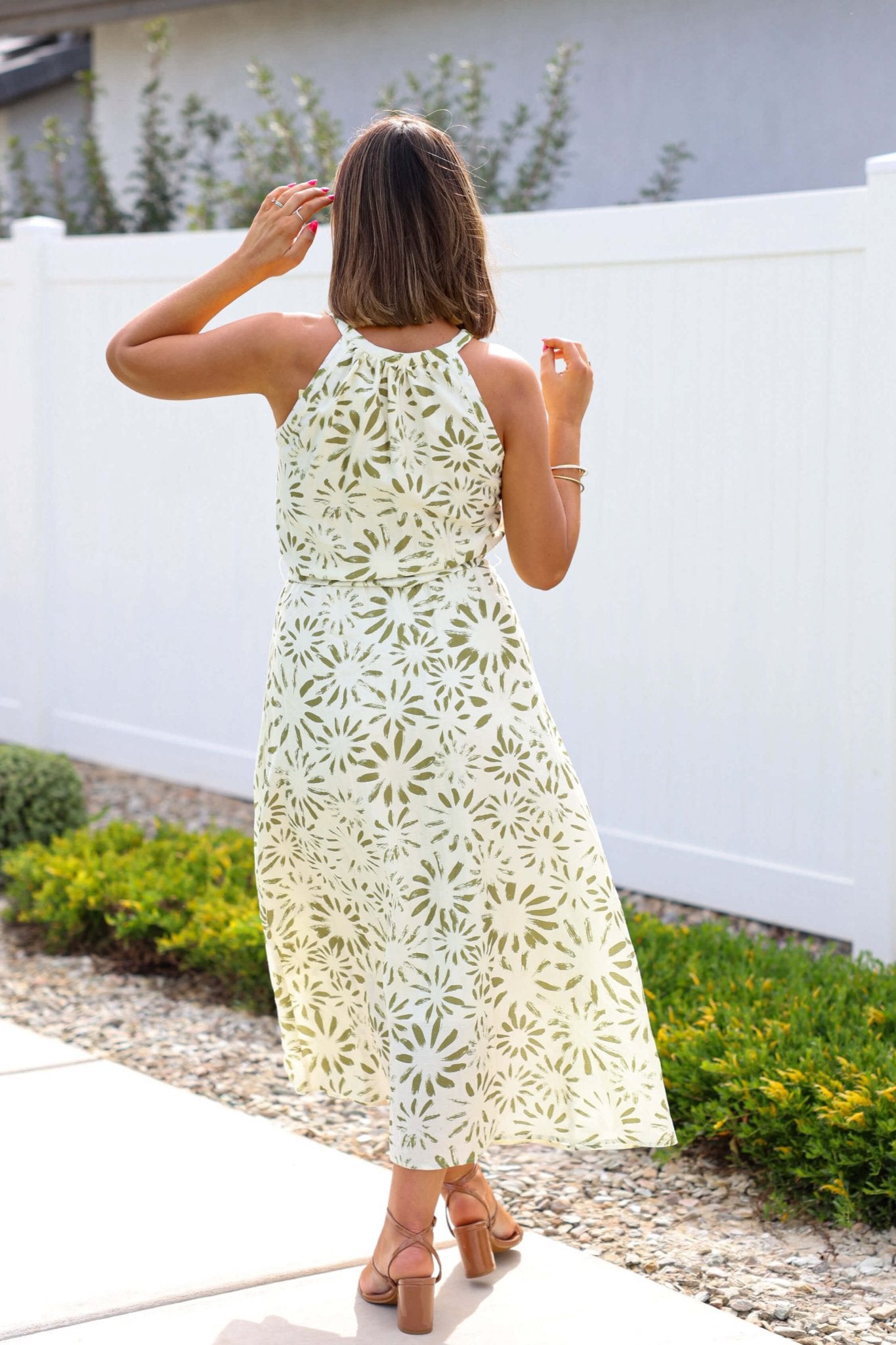 Woman wearing the Graceful Olive Floral Print Belted Midi Dress and sandals stands by a white fence, facing away.