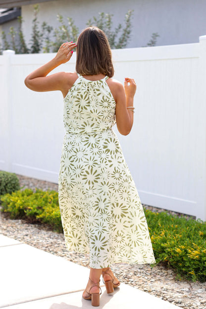 Woman wearing the Graceful Olive Floral Print Belted Midi Dress and sandals stands by a white fence, facing away.