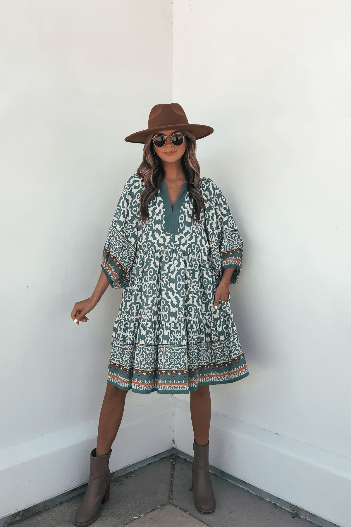 Woman in a Green Border Print Tiered Mini Dress poses confidently against a white wall, accessorized with a brown hat and sunglasses.
