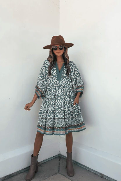 Woman in a Green Border Print Tiered Mini Dress poses confidently against a white wall, accessorized with a brown hat and sunglasses.