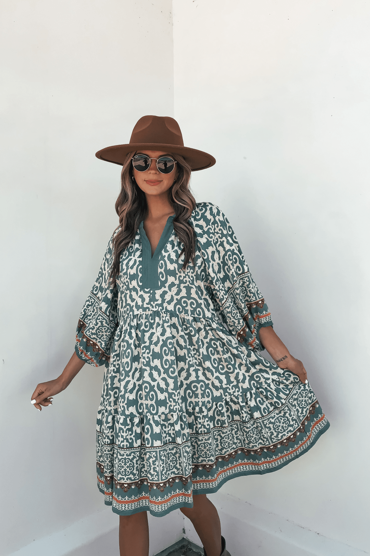Woman poses in a Green Border Print Tiered Mini Dress - FINAL SALE, styled with a brown hat and sunglasses against a white wall.