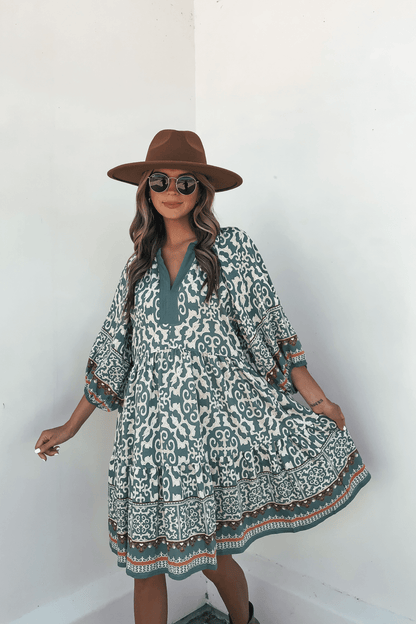 Woman poses in a Green Border Print Tiered Mini Dress - FINAL SALE, styled with a brown hat and sunglasses against a white wall.