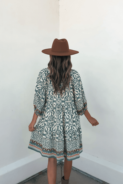 Woman in a Green Border Print Tiered Mini Dress - FINAL SALE and brown hat stands with her back to the camera by a white wall.