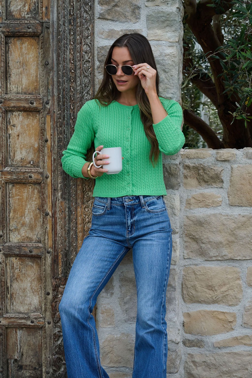 A woman in a Green Cable Knit Sweater Cardigan and blue jeans holds a white mug, adjusting sunglasses by a rustic stone wall.