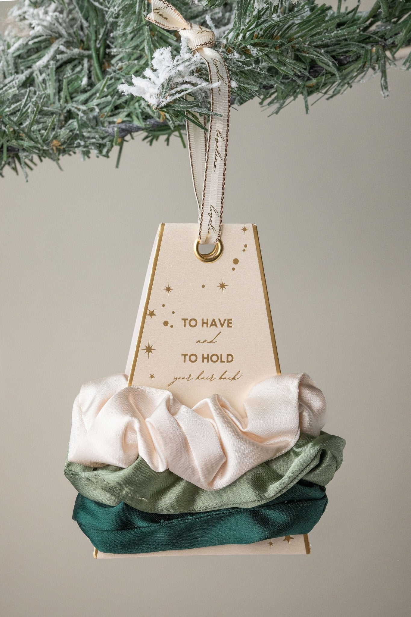 The Green Gaja Satin Scrunchie Set in cream, sage, and dark green is a perfect gift, beautifully displayed on a snowy pine branch.