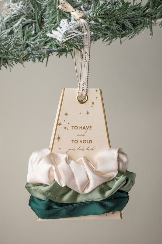 The Green Gaja Satin Scrunchie Set in cream, sage, and dark green is a perfect gift, beautifully displayed on a snowy pine branch.