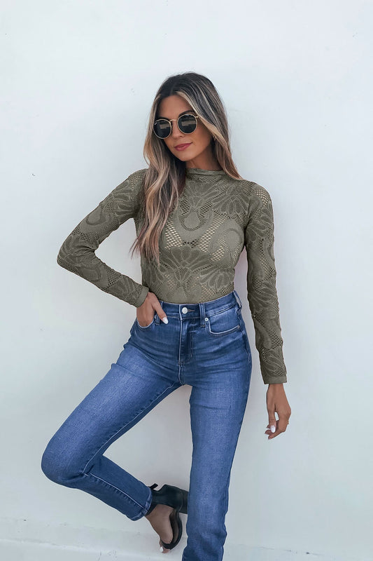 Woman in sunglasses wears a Green Mock Neck Fishnet Top and blue jeans, posing with one leg crossed against a white wall.