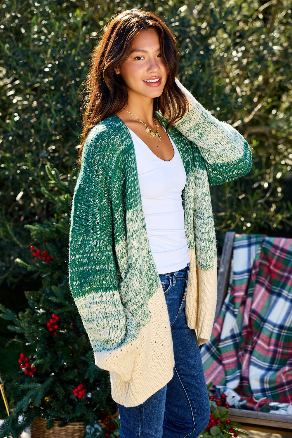 A woman wears the Green Ombre Open Front Cardigan - FINAL SALE with a white top and jeans by holiday greenery and a plaid blanket.