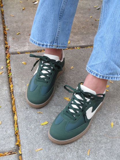 Person in light blue jeans and Green Remus Lace Up Sneakers - DOORBUSTER, standing on a sidewalk with scattered yellow leaves.