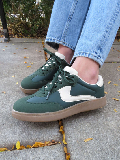 Person in Green Remus Lace Up Sneakers - DOORBUSTER and light blue jeans stands on a sidewalk scattered with yellow leaves.