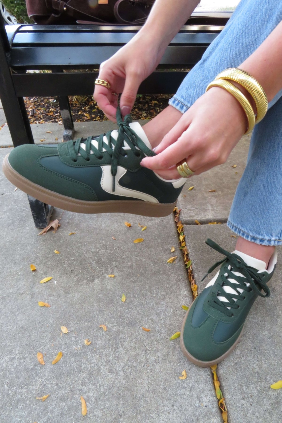 Someone in jeans and gold jewelry ties the laces of their Green Remus Lace Up Sneakers - DOORBUSTER while on a bench.