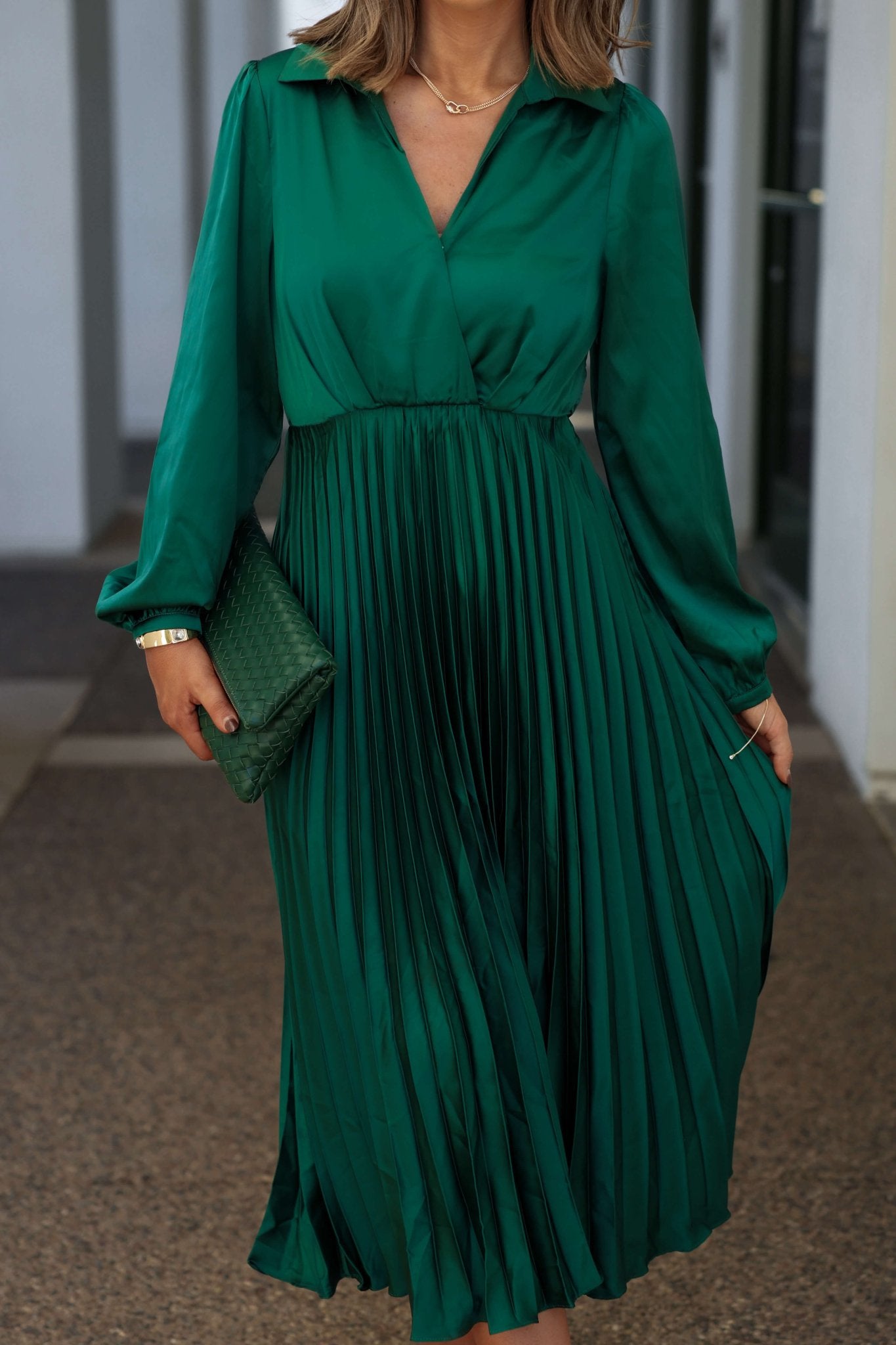 A woman stands outdoors in a Green Satin Pleated Midi Dress, holding a matching clutch.