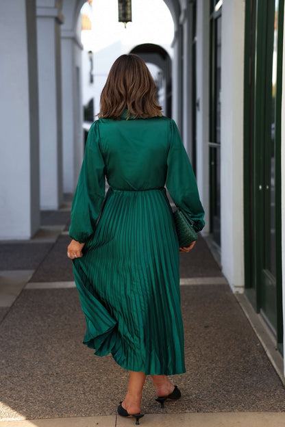 A woman walks away down a covered outdoor corridor in a Green Satin Pleated Midi Dress and black heels.